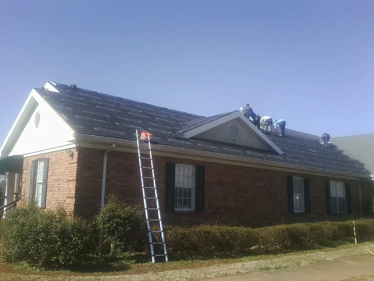 Crew tearing shingles off a brick ranch roof with a ladder set — Emergency Roof Repair work by our crew in Mustang