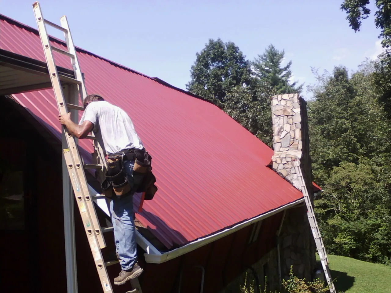 Roofer on a ladder beside a red metal roof and stone chimney — Soffit & Fascia Repair by Roofing Brothers in Mustang